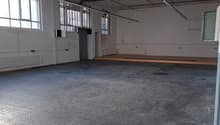 Large empty warehouse room, white walls, metal flooring, fluorescent ceiling lights, windows, radiators, small raised platform, door