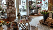 Room filled with decorative items and floral arrangements, white tiled flooring, wooden tables and benches, natural light from windows