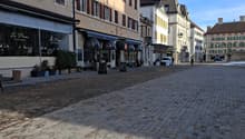 Cobblestone street with buildings on both sides, shops, trees, street lamps, people walking, cars parked.