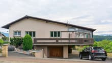 2-story house with stucco exterior, brown roof, large windows, wooden garage door, balcony, and a parked car in the driveway. The house is surrounded by landscaping with shrubs and trees.