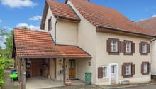 Two-story house with red roof, beige walls, wooden windows, attached garage, brick driveway.