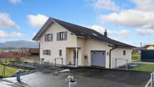 Modern house, beige exterior, black roof, large windows, two garages, driveway, fenced garden, mountains in the distance