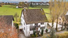 A white house with a balcony, brown roof, situated in a village, surrounded by green fields and mountains.