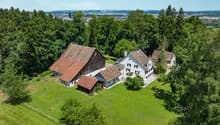 Two-story farmhouse with a red tile roof, surrounded by lush green trees and a large grassy lawn. The property also includes a smaller wooden barn-like structure. The houses have white walls and windows, and there appears to be a balcony or terrace on one