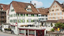 A multi-story building with a traditional European architectural style, featuring red-tiled roofs, green shuttered windows, and a balcony. The building houses a tea room on the ground floor, with outdoor seating. The surrounding buildings have a similar s