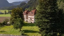 Large historic manor house with red-tiled roofs surrounded by lush green forests and mountains in the background. The house appears to be situated on a grassy field with a stone wall in the foreground.
