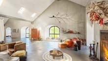 Spacious open-concept living area with high ceilings, arched windows, concrete walls, and a fireplace. The room features a mix of rustic and modern decor elements, including a large orange sofa, wicker furniture, and decorative accents like a hanging plan