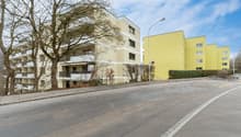 Large multi-story building with balconies, beige exterior, modern design, surrounded by trees and bushes, clear sky