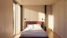 A bedroom with a large bed, white walls, wooden floor, large windows with white curtains, and two wall lamps