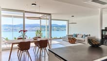 Spacious open-plan living and dining area with large windows offering a scenic view of the lake and surrounding mountains. The dining table with wooden chairs is situated near the windows, while the living room features a comfortable sofa and armchairs. T