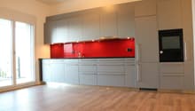 Modern kitchen with gray cabinets, red backsplash, stainless steel faucet, black oven, and wooden floors.