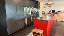 Modern kitchen, gray cabinets, red island counter, wooden step stool, wooden cabinets on the right side, stainless steel range hood, tiled floor.