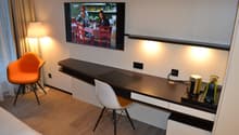 Modern living room with a large TV mounted on the wall, a dark wooden TV stand with drawers, an orange accent chair, and a white chair at a desk area. The room has hardwood flooring and appears to have a balcony or terrace visible through the window.