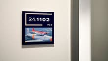 The image shows a framed display with the number 34.1102 and the label 'PC-3'. The display also includes a photograph of a red and white aircraft, likely a military jet or trainer aircraft.