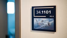 The image shows a room with a wall-mounted plaque displaying the number 34.11101 and the text 'PC-2', along with an image of an aircraft. The room has a white wall and a blue-tinted window.