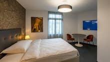 Spacious bedroom with a large bed, leather headboard, and a seating area with a round table and two chairs. The room has large windows with curtains, and a flat-screen TV on the wall. There is also a framed artwork on the wall and a ceiling light fixture.