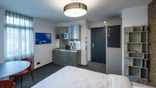 The image shows a modern studio apartment with a kitchenette, a bed, and a living area. The kitchenette has a microwave, a sink, and a dishwasher. The living area has a TV mounted on the wall and a small dining table with chairs. The room has large window