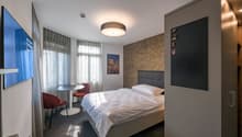 A bedroom with a single bed, bedside tables, a TV, a painting on the wall, and two red chairs next to a round table.