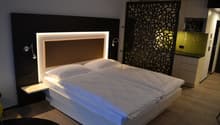 Modern bedroom with a large bed, white bedding, and a backlit headboard. The room also features a decorative screen on the wall and a kitchenette area.