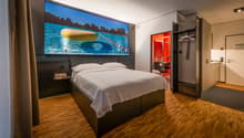 Large bedroom with a king-size bed, hardwood flooring, and a large wall-mounted TV displaying a scenic image of a lake and mountains. The room also has a closet and a bathroom with a red accent wall.