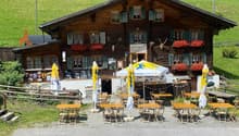 Traditional Swiss chalet-style building with wooden exterior, decorated with flower boxes and antlers. Outdoor seating area with yellow umbrellas and tables. Appears to be a restaurant or cafe in a rural, mountainous setting.