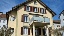 3 story beige house with brown shutters, glass balcony on the second floor, beige pillars, brown roof