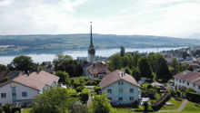 Aerial view of a picturesque town nestled among lush greenery, with a church steeple prominently visible in the center. The town is situated near a large body of water, likely a lake or river, with rolling hills and mountains in the background.