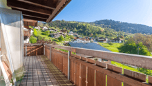 Wooden balcony with a scenic view of a mountainous village with houses, trees, and a clear blue sky.