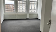 This image shows an empty room with large windows overlooking a city skyline. The room has white walls, a grey carpeted floor, and a track lighting fixture on the ceiling. The room appears to be a living or office space.