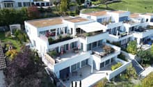 Modern white apartment building, 3 stories, multiple balconies, terrace area with umbrellas, garden areas, view of the surrounding countryside.