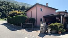 Two-story pink house with a tiled roof, surrounded by trees and greenery. The house has a garage and a stone wall with a gate in the front. There are potted plants and other decorative elements around the entrance.