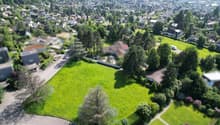 An aerial view of a suburban neighborhood with houses, green fields, and trees. The houses are mostly detached single-family homes with well-kept yards. The area is surrounded by trees, and there are hills in the background. A driveway and car are visible