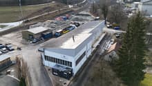 Industrial building with several trucks parked, storage area with containers and a small building.