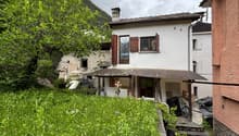 Two-story house with white walls, red roof tiles, and wooden shutters. The house is surrounded by lush greenery, including trees and plants. There is a small balcony or terrace visible on the upper level. The house is situated in a mountainous, rural area