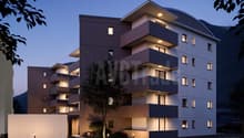 Multi-story apartment building with modern, angular design featuring balconies and terraces, outdoor lighting, and a parking area in the foreground