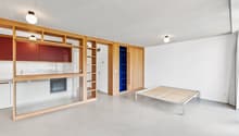 Open-concept studio apartment with wooden shelving units, a kitchenette with a dishwasher, and a raised platform bed area