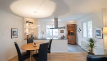 The image shows an open concept living space with a kitchen, dining area, and living room. The kitchen features a black range hood and built-in appliances. The dining area has a round wooden table with black chairs. The living room has a wooden cabinet an