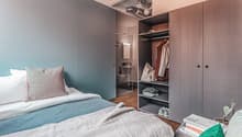 Bedroom with double bed, open door, wooden floor, wardrobe with clothes, bedside table with potted plant, and open bathroom with toilet.