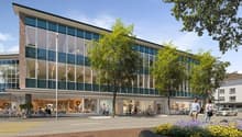 A modern, multi-story commercial building with large glass windows and a prominent entrance. The building has retail stores on the ground floor and appears to be located in a pedestrian-friendly urban area with trees, flowers, and people walking around.