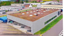 Large commercial building with a green roof, surrounded by parking spaces and other industrial buildings. The building has a modern, industrial design with a flat roof and large windows.