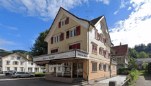 3-story building, brick facade, red shutters, storefront at ground level, small car park in front, green trees, neighboring buildings visible in background