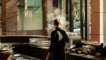 The image shows the interior of a restaurant kitchen. A chef in a black uniform is standing in front of a large commercial kitchen appliance, with steam or smoke visible. The kitchen appears to be well-equipped with various cooking utensils, dishes, and g