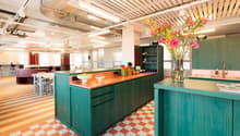 Open concept office kitchen with colorful, modern design. Features a large central island with wooden countertop, green cabinets, and a dishwasher. Bright lighting, checkered tile flooring, and decorative floral arrangement add to the vibrant atmosphere.