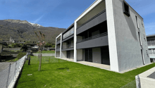 3-story modern apartment building with large windows, balconies, and a grassy lawn in front surrounded by a fence