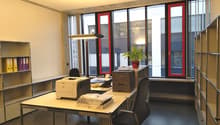 The image shows an office space with a desk, chair, printer, and shelving units. The room has large windows with red frames, providing natural light. There is a potted plant on the desk, adding a touch of greenery to the space.