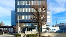 A multi-story office building with a modern glass and steel facade. The building has several floors and windows, and there is a tree in front of the entrance. The building appears to be located in an urban area with other buildings and vehicles visible in