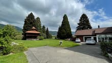 House exterior, parking area, two cars parked, wooden barn, lush green lawn, trees, mountainous background