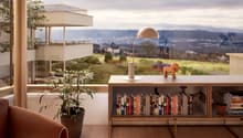 Spacious living room with large windows overlooking a scenic outdoor landscape, including a balcony or terrace. The room features a wooden bookshelf with various books, a potted plant, and a decorative figurine. The overall design has a modern and cozy fe
