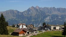 The image shows a picturesque mountain town nestled in a valley surrounded by rugged, snow-capped peaks. The town features a mix of traditional wooden chalets and modern buildings, with a parking area visible in the foreground. The landscape is dominated
