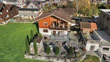 Two-story wooden chalet-style house with a balcony, surrounded by a landscaped garden with trees and shrubs. The house is situated in a residential area with other houses and buildings visible in the background.